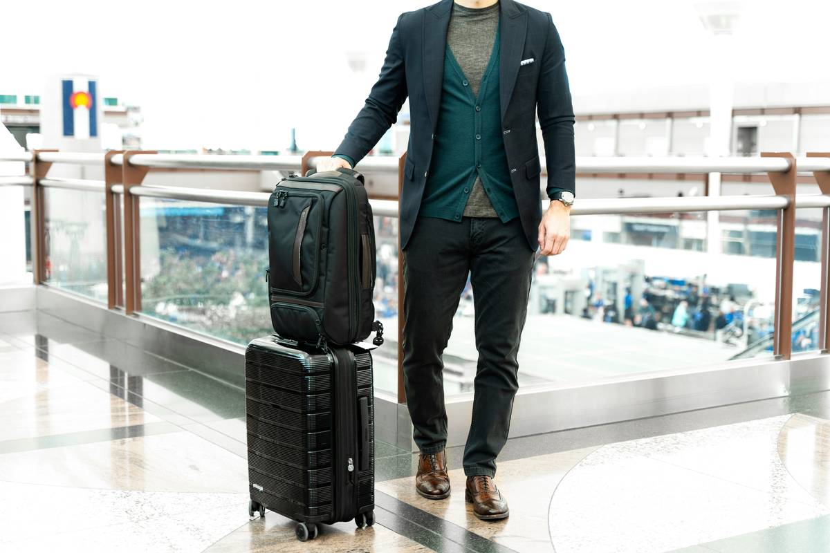 Black armored travel bag case with spinner wheels on airport tarmac, showing scuff marks but intact structure